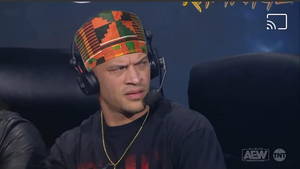 A photo of Ricky Starks sitting at the commentary desk with a kufi photoshopped onto him. He's looking off to the side (presumably at someone) with a puzzled expression.