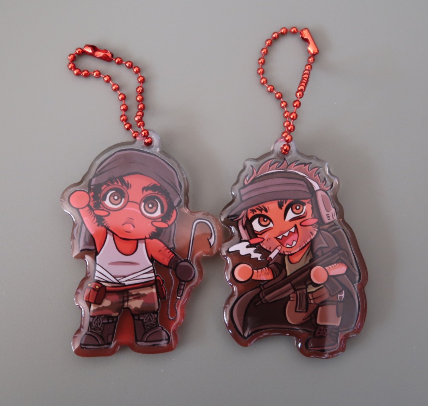 A photo of the two Sanmos charms, featuring my chibi designs, a red gradient background, and a red ball chain connector.